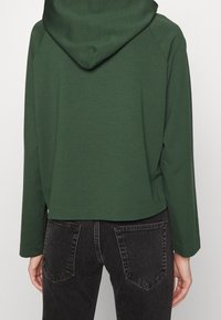 Felpa verde corta con cappuccio, caratterizzata da maniche lunghe raglan. Il materiale sembra morbido con una texture liscia. Abbinata a jeans scuri.