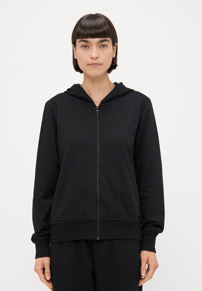 Schwarze Zip-up Kapuzenjacke mit einer Fronttasche, gerippten Bündchen und gesticktem Logo auf der linken Brust. Weicher Stoff mit einer glatten Textur.
