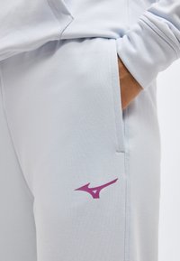 Χέρι ατόμου στην τσέπη ανοιχτόχρωμων γκρι παντελονιών φόρμας με μικρό μωβ λογότυπο αθλητικού εμπορικού σήματος στον μηρό.