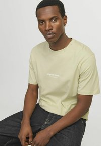 Limonengrünes Baumwoll-T-Shirt mit Rundhalsausschnitt, kurzen Ärmeln und weißem "Originals Studio" Grafikdruck auf der Brust. Getragen mit dunklen Jeans.