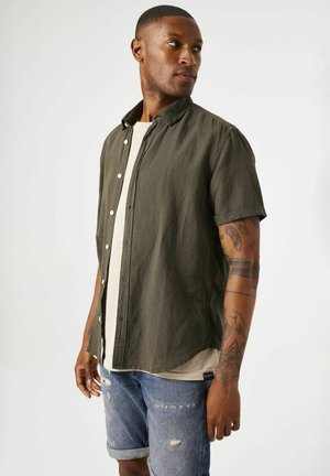 Camisa de manga corta, con botones, en tejido verde oliva oscuro, con un corte relajado y un dobladillo ligeramente curvado, llevada sobre una camiseta de color claro.