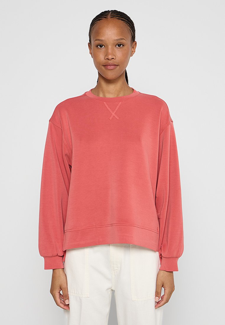 Junge Person mit geflochtenen Haaren, die einen lockeren korallenfarbenen Sweatshirt und weiße Hosen trägt, vor einem schlichten hellen Hintergrund steht.