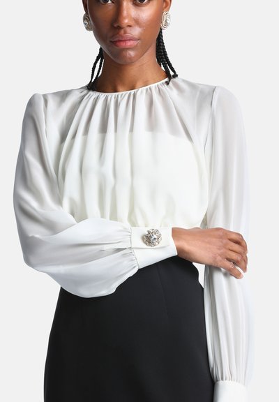 Femme portant un chemisier blanc à manches longues avec un col froncé et des boutons bijoux aux poignets, associé à une jupe ou un pantalon noir.