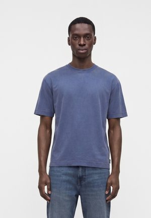 Uomo in piedi contro uno sfondo semplice, indossa una maglietta blu a maniche corte e jeans blu, rivolto in avanti con le braccia rilassate lungo i fianchi.