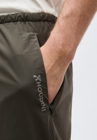 Gros plan d'une main dans la poche d'un pantalon vert olive avec ceinture élastique et une poche latérale zippée visible.