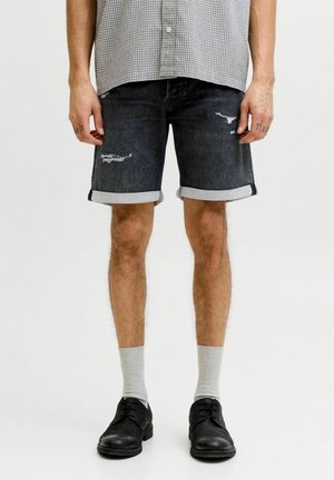 Mężczyzna noszący podwinięte czarne szorty z denimu z przetarciami, jasnoszare skarpetki, czarne buty oraz szarą koszulę w kratę.