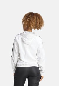 Witte gevoerde jas met elastische tailleband, voorzien van zwarte zijstrepen. De stof lijkt licht en glanzend. Gedragen boven zwarte legging.