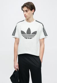 Fehér pamut póló fekete grafikákkal és három fekete csíkkal minden vállon. A kiemelt logó egy stilizált levélminta. Fekete nadrág van rajta.