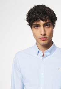 Hellblaues Button-down-Hemd aus Baumwolle, mit einem spitzen Kragen, langen Ärmeln und einem kleinen gestickten Logo auf der Brust.