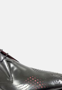 Zapato de cuero negro con detalles perforados y acentos rojos, con una forma elegante y cordones, diseñado para vestir formalmente.