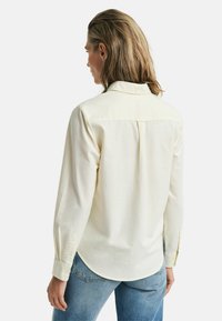 Light yellow button-up shirt with long sleeves, a back yoke, and rounded hem. Fabric appears soft with a subtle texture. Worn with blue jeans.