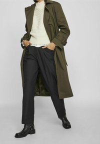 Oliwkowy trencz na białym swetrze w warkocze, czarne spodnie i czarne botki na kostkę z dodatkowymi paskami, stojący na neutralnym tle.