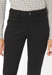 Frau, die hoch taillierte schwarze Hose mit Fronttaschen und Knopfverschluss trägt, kombiniert mit einem weißen gemusterten Langarmoberteil.