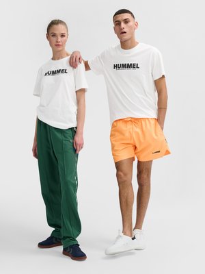 Vrouw in wit Hummel T-shirt, groene trainingsbroek en marineblauwe schoenen; man in wit Hummel T-shirt, oranje korte broek en witte sneakers, staat nonchalant.