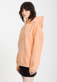 Hoodie w kolorze brzoskwiniowym z kieszenią kangurzą, kapturem ze sznurkiem i ściągaczami przy mankietach. Gładka faktura i luźny fason.