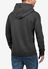 Sudadera gris oscuro con capucha, puños y dobladillo acanalados. Presenta una pequeña etiqueta con el logo en la parte inferior izquierda. Se lleva con jeans azules.