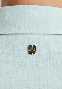 Hellblaue Leinenbluse mit einem schwarzen und goldenen PME Legend-Logoschild, runder Kragen und subtiler Textur.