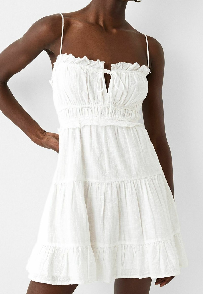 Mörkhyad person klädd i en vit ärmlös miniklänning med volangmidja, tunna axelband och lager av kjol, poserar med handen på höften.