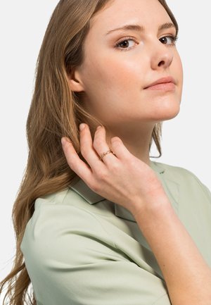Bague dorée avec un design en maillons de chaîne portée à un doigt, associée à une blouse à col vert clair et de longs cheveux ondulés.