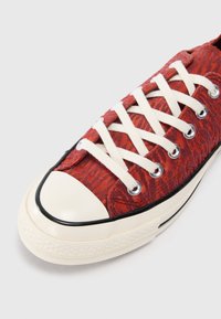 Rød mønstrete lærredssko med hvite snøringer, svart gummitåhette og teksturert stoffdetalj. Hvit gummisåle med sømmer.