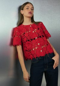 Sarkanais mežģīņu blūze ar ziedu izgriezumu, īsām piedurknēm un zelta pogu aizdarēm. Stilizēta ar tumšas džinsu bikses.