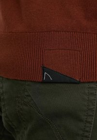 Brązowy dziergany sweter z prążkowanym zakończeniem, ozdobiony małą czarną tkaninową naszywką z białym wzorem, zestawiony z zielonymi spodniami z dekoracyjnym przeszyciem.