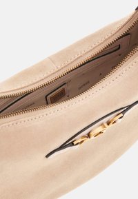 Beige Wildledertasche mit einem goldfarbenen Reißverschluss, das über eine strukturierte Außenseite, schwarze Akzente und ein Innenfutter mit Logo-Muster verfügt.