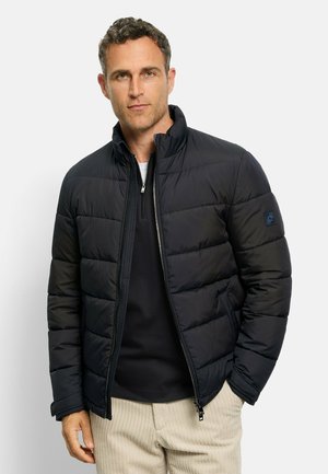 Schwarze Steppjacke mit Reißverschluss vorne, hohem Kragen und gestepptem Design. Verfügt über ein kleines Logo auf dem linken Ärmel. Getragen über einem schwarzen Oberteil.
