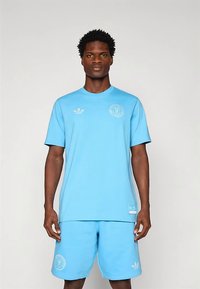 Helehall sinine spordikomplekt, mis sisaldab ümar kaelusega lühikeste varrukatega t-särki ja sobivaid lühikesi pükse, valmistatud pehmest kangast ja logodetailidega.