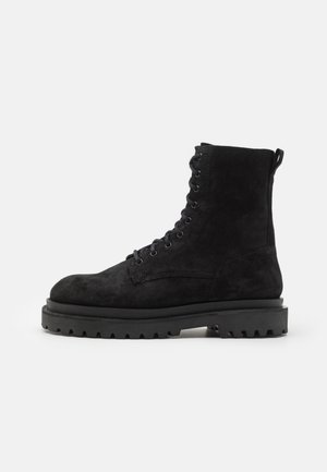 Bottines de combat en suede noir à lacets avec semelle en caoutchouc épaisse et boucle au talon, présentées de profil sur un fond blanc.