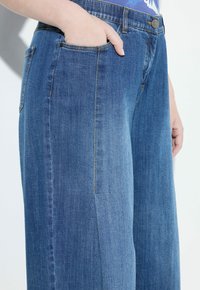 Mano en el bolsillo de unos jeans de mezclilla azul medio, combinados con una blusa floral azul metida por dentro, sobre un fondo blanco.