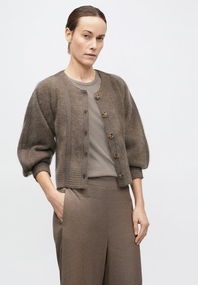 Femeie purtând un cardigan maro pufos cu nasturi și mâneci bufante peste un top maro și pantaloni largi cu talie înaltă asortați.