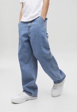 Henkilö, jolla on päällään väljät siniset farkut, valkoiset tennarit ja valkoinen t-paita, kädet taskuissa, seisoo yksinkertaisen taustan edessä.