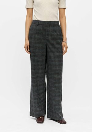 Femme portant un pantalon à taille haute, jambes larges, à carreaux gris foncé, associé à des chaussures pointues marron et un top beige à manches courtes.
