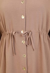 Haut en tissu texturé marron clair avec une taille cintrée, comprenant des boutons dorés sur le devant et des liens à la taille pour un ajustement réglable.