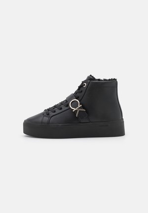 Zapatilla negra de caña alta con cordones delanteros, suela gruesa, forro de piel de borrego y una hebilla plateada con el logo "OK" en la correa lateral.