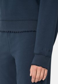 Sada námořnické modré mikiny a kalhot vyrobených z měkké tkaniny, s žebrovanými manžetami a roztrhanými lemy. Obsahuje embosované logo.