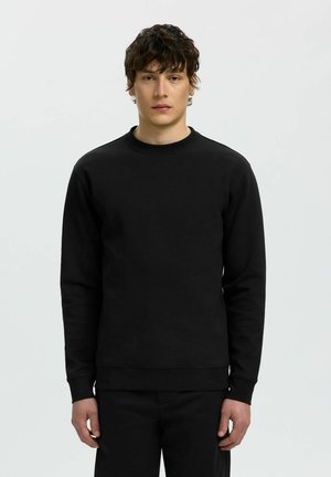 Ung mand med kort krøllet mørkt hår iført en ensfarvet sort langærmet sweatshirt og sorte bukser mod en lys baggrund.