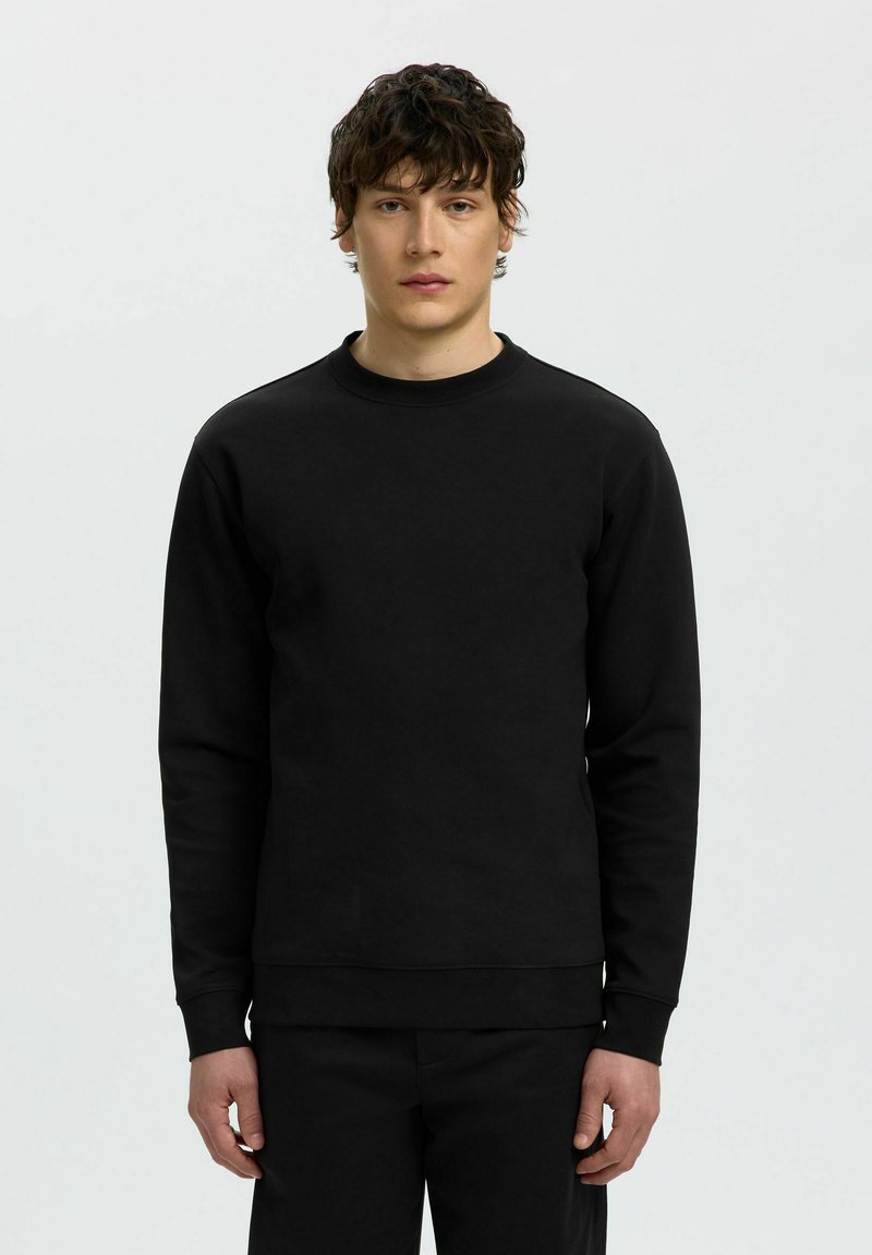 Jeune homme aux cheveux courts, bouclés et foncés, debout, portant un sweat-shirt noir uni à manches longues et un pantalon noir sur un fond clair.