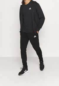 Schwarzes Sportoutfit mit einem Kapuzenpullover mit Reißverschluss, einem kurzärmeligen Shirt und Jogginghosen, alle mit weißen Adidas-Logo-Akzenten. Schwarze Sneakers runden den Look ab.