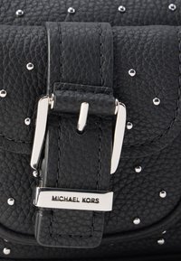 Τσάντα χειρός από μαύρο δερμάτινο υλικό με υφή, διακοσμημένη με ασημένιες τρουκ κ αι μεταλλική αγκράφα με πλακέτα που φέρει το όνομα της μάρκας. Κοντινή λήψη της λεπτομέρειας του κλεισίματος.