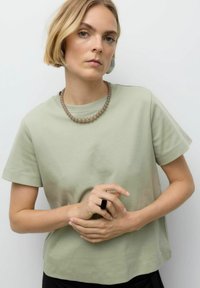 Lys grøn bomulds T-shirt med korte ærmer. Modellen bærer store statement øreringe og en perlekæde, mens hun holder hænderne samlet. Sort ring accent.