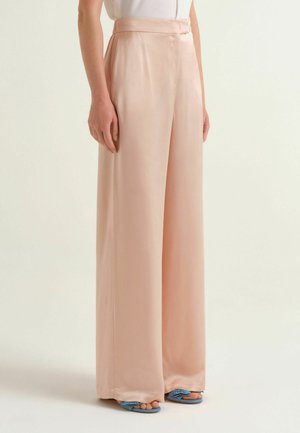 Femme portant un pantalon pêche taille haute à jambes larges, un haut blanc et des talons bleus ouverts à l'avant, debout devant un fond clair uni.