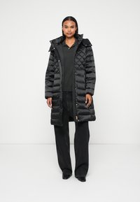 Casaco puffer preto com um design acolchoado, capuz e detalhes em dourado, combinado com jeans pretos largos e uma blusa escura.