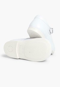 Witte schoenen met een glanzende afwerking, voorzien van een gesp aan de zijkant, en met gestructureerde witte zolen met een gepatternde grip.