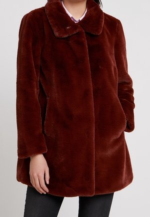 Femme portant un manteau en fausse fourrure brun foncé et pelucheux avec un large col, des poches avant et des manches longues, par-dessus un pantalon noir.