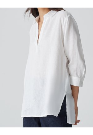 Kobieta ubrana w luźną białą lnianą tunikę z rękawami do łokcia i bocznymi rozcięciami, zestawioną z ciemnoniebieskimi jeansami.