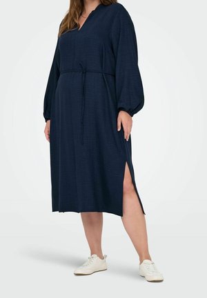 Woman wearing a long-sleeve navy midi dress with waist tie and side slit, paired with white sneakers, standing against white background.