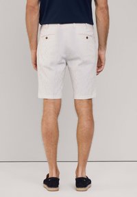 Pantaloni corti a righe bianche con una vestibilità sartoriale, dotati di tasche posteriori con bottoni marroni e una texture liscia. Indossati con scarpe slip-on scure.