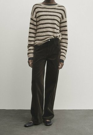 Person wearing a beige and brown striped long-sleeve sweater, dark wide-leg pants, and black slip-on shoes standing on a gray floor.